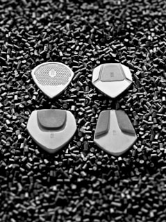 Four silver guitar picks are placed on a bed of small black cylindrical pellets. The picks are arranged in a neat pattern, showcasing various textures and designs on each.