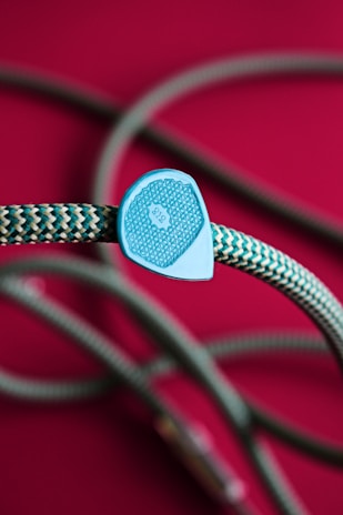 A braided rope with a zigzag pattern in shades of teal and white, accented by a plastic piece with intricate designs and the number 9.8. The background is a rich magenta, which contrasts with the color of the rope.