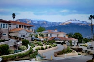 A picturesque residential area features large, upscale houses with red-tiled roofs set against a backdrop of snow-capped mountains. The landscape includes meticulously designed gardens with various shrubs and palm trees. The environment appears tranquil and well-maintained, with a clear view of the scenic mountain range under a partly cloudy sky.