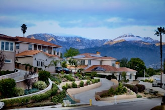 A picturesque residential area features large, upscale houses with red-tiled roofs set against a backdrop of snow-capped mountains. The landscape includes meticulously designed gardens with various shrubs and palm trees. The environment appears tranquil and well-maintained, with a clear view of the scenic mountain range under a partly cloudy sky.