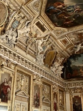 An opulently decorated room featuring intricate golden moldings and detailed paintings on the ceiling and walls. Numerous sculptures adorn the space, including figures seemingly in motion. Portraits in ornate frames line the walls, adding to the luxurious atmosphere.