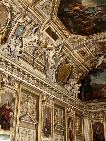 An opulently decorated room featuring intricate golden moldings and detailed paintings on the ceiling and walls. Numerous sculptures adorn the space, including figures seemingly in motion. Portraits in ornate frames line the walls, adding to the luxurious atmosphere.