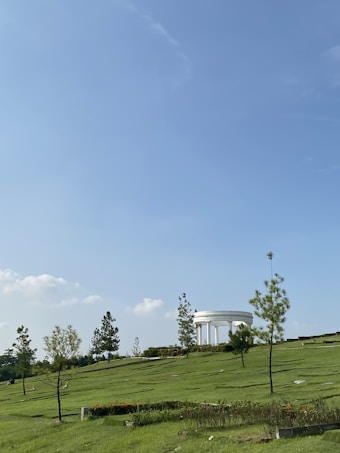 A grassy landscape with gently sloping hills and scattered trees leads up to a white, circular structure with columns in the background. The sky is clear with a few fluffy clouds, creating a serene, peaceful setting.