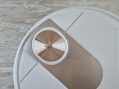 A modern wall clock with a minimalist design features a circular face and metallic hands. The clock is positioned against a wooden textured background, giving it a contemporary yet natural feel.