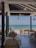 An open-air restaurant by the seaside includes several dining tables with neatly arranged white tablecloths and minimalistic table settings. The restaurant has a wooden floor and a partially covered ceiling that shelters the tables. Potted plants and small thatched umbrellas can be seen outside on the beach providing shade. The clear blue ocean water and a few boats are visible in the background, under a bright, slightly cloudy sky.