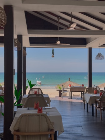 An open-air restaurant by the seaside includes several dining tables with neatly arranged white tablecloths and minimalistic table settings. The restaurant has a wooden floor and a partially covered ceiling that shelters the tables. Potted plants and small thatched umbrellas can be seen outside on the beach providing shade. The clear blue ocean water and a few boats are visible in the background, under a bright, slightly cloudy sky.
