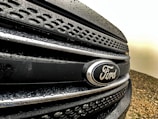 Close-up view of a Ford vehicle grille with water droplets on a dark surface, showcasing the Ford emblem prominently.