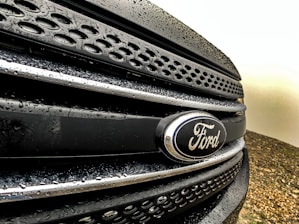 Close-up view of a Ford vehicle grille with water droplets on a dark surface, showcasing the Ford emblem prominently.