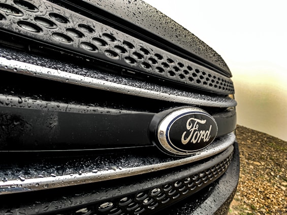 Close-up view of a Ford vehicle grille with water droplets on a dark surface, showcasing the Ford emblem prominently.
