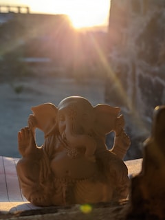 A small statue of a Hindu deity with an elephant head is placed on a surface, partially illuminated by the warm glow of the setting sun. The statue features intricate details in its carving, capturing the essence of traditional craftsmanship. The sunlight creates a serene atmosphere, casting soft shadows.