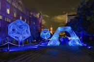 A night scene featuring large, geometric light installations in vibrant blue placed along a pathway beside a historic brick building. The structures exhibit intricate linear patterns and are illuminated, casting a glow on the surrounding area. Several people are walking underneath a large, arch-shaped light installation.