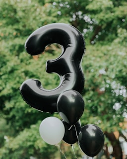 A large, black balloon shaped like the number three is surrounded by several smaller balloons, including white and black ones, floating against a blurred background of lush green leaves.