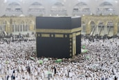 A large crowd of people dressed in white surround and walk around the Kaaba, an enormous black cube structure adorned with gold calligraphy. The environment is bustling with people, some holding umbrellas, which suggests it may be raining. The background shows grand architectural arches and a two-story gallery filled with observers.