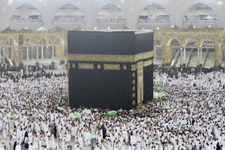 A large crowd of people dressed in white surround and walk around the Kaaba, an enormous black cube structure adorned with gold calligraphy. The environment is bustling with people, some holding umbrellas, which suggests it may be raining. The background shows grand architectural arches and a two-story gallery filled with observers.