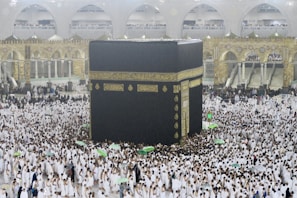 A large crowd of people dressed in white surround and walk around the Kaaba, an enormous black cube structure adorned with gold calligraphy. The environment is bustling with people, some holding umbrellas, which suggests it may be raining. The background shows grand architectural arches and a two-story gallery filled with observers.