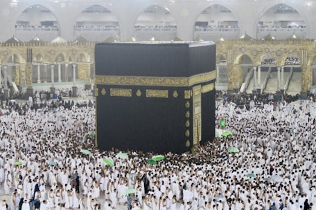 A large crowd of people dressed in white surround and walk around the Kaaba, an enormous black cube structure adorned with gold calligraphy. The environment is bustling with people, some holding umbrellas, which suggests it may be raining. The background shows grand architectural arches and a two-story gallery filled with observers.