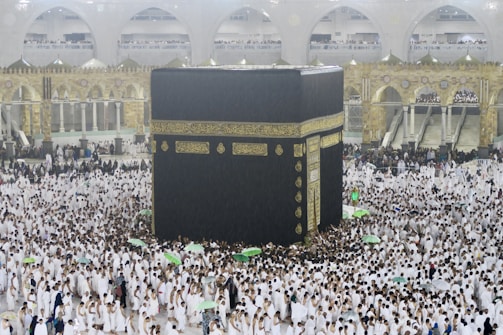 A large crowd of people dressed in white surround and walk around the Kaaba, an enormous black cube structure adorned with gold calligraphy. The environment is bustling with people, some holding umbrellas, which suggests it may be raining. The background shows grand architectural arches and a two-story gallery filled with observers.