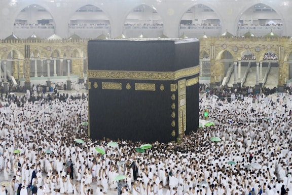 A large crowd of people dressed in white surround and walk around the Kaaba, an enormous black cube structure adorned with gold calligraphy. The environment is bustling with people, some holding umbrellas, which suggests it may be raining. The background shows grand architectural arches and a two-story gallery filled with observers.