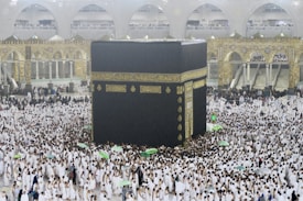 A large crowd of people dressed in white surround and walk around the Kaaba, an enormous black cube structure adorned with gold calligraphy. The environment is bustling with people, some holding umbrellas, which suggests it may be raining. The background shows grand architectural arches and a two-story gallery filled with observers.