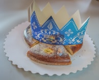 A sweet, circular pastry is placed on a scalloped white paper tray, topped with sugar. A decorative blue and white paper crown rests on top, adorned with ornate patterns. A small figurine resembling a pig is situated in front of one of the cut pieces of pastry.