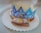 A sweet, circular pastry is placed on a scalloped white paper tray, topped with sugar. A decorative blue and white paper crown rests on top, adorned with ornate patterns. A small figurine resembling a pig is situated in front of one of the cut pieces of pastry.