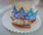 A sweet, circular pastry is placed on a scalloped white paper tray, topped with sugar. A decorative blue and white paper crown rests on top, adorned with ornate patterns. A small figurine resembling a pig is situated in front of one of the cut pieces of pastry.