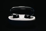 A pair of black wireless earbuds nestled within a white charging case with a translucent lid. The background is very dark, which highlights the earbuds and the case in the foreground.