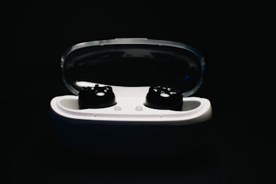 A pair of black wireless earbuds nestled within a white charging case with a translucent lid. The background is very dark, which highlights the earbuds and the case in the foreground.