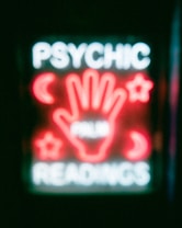 A neon sign featuring the words 'Psychic Readings' with a central image of a hand surrounded by symbols like crescent moons and stars.