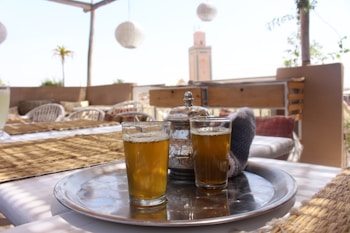 A rooftop setting with a silver tray holding two glasses of tea and a teapot. The table is covered with woven mats, and wicker chairs are around it. In the background, a tall tower-like structure and a palm tree are visible under a clear blue sky, contributing to a relaxed and exotic atmosphere.