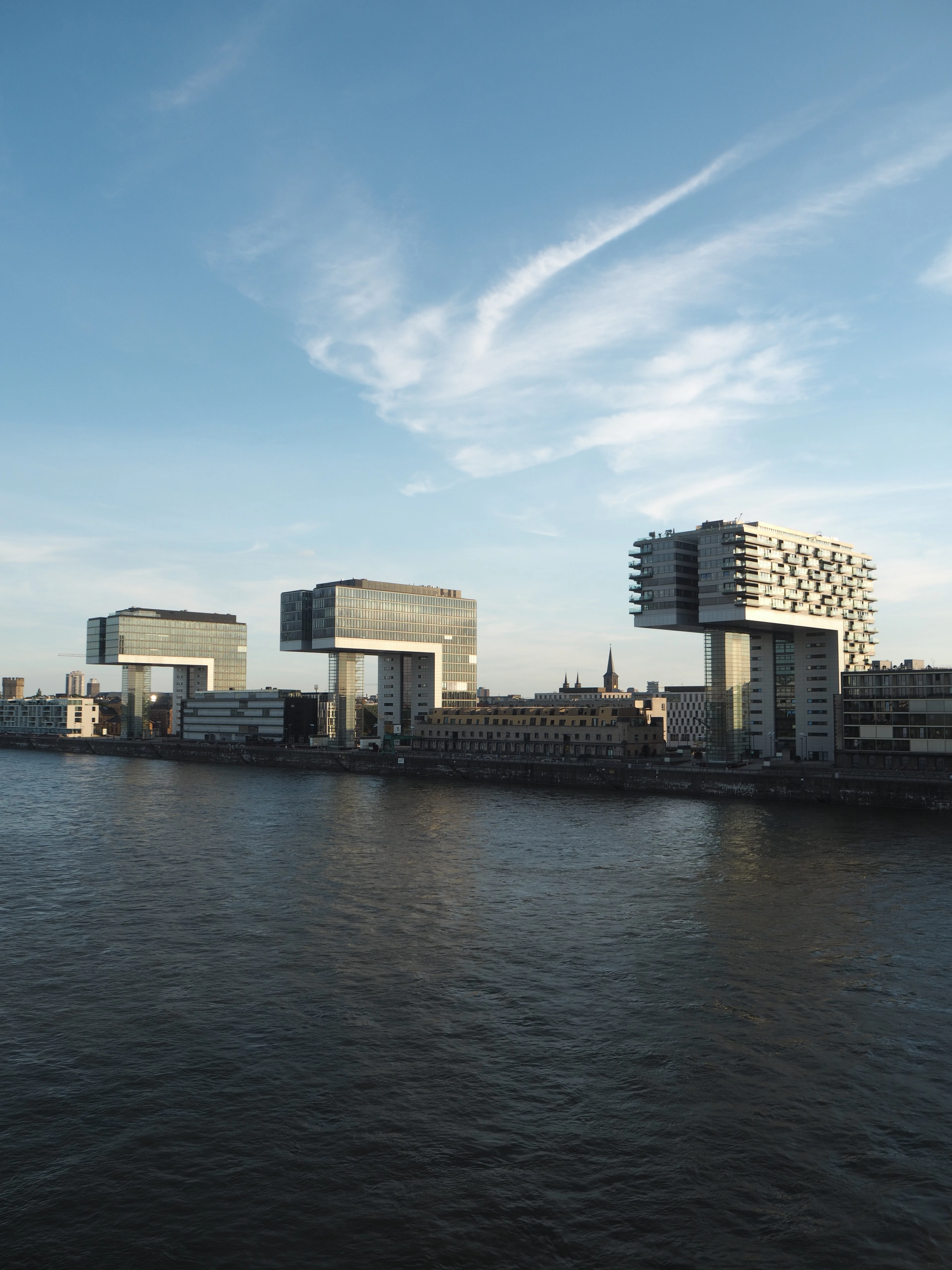 Three Kranhaus crane-shaped buildings along the Rhine in Cologne under a blue sky with wispy clouds.