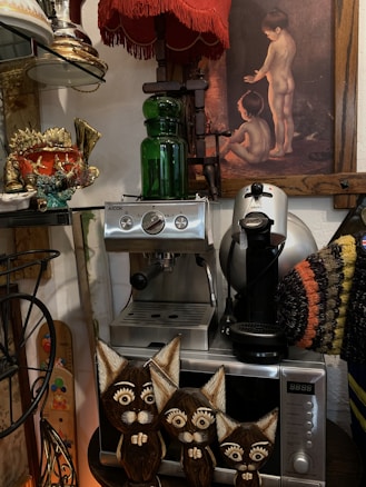 A collection of eclectic items including an espresso machine, a painting of two children, a colorful ceramic fish sculpture, a green glass bottle, and three wooden cat figurines. There is also a vibrant red lampshade, a coffee maker, and a knit item with striped patterns, all arranged on a shelf.