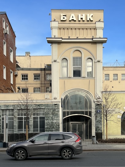 A tall building with a classical architectural design featuring large arched windows and decorative elements. The sign at the top reads 'BANK' in Cyrillic script. There is a parked SUV on the street in front of the building, with surrounding structures displaying a mix of brick and concrete facades.