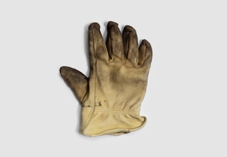A single worn-out, dirty work glove with a leather-like texture lies against a plain background. The glove has visible signs of use with dark stains and scuffs on the fingertips and palm.