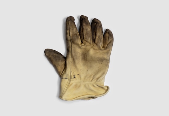 A single worn-out, dirty work glove with a leather-like texture lies against a plain background. The glove has visible signs of use with dark stains and scuffs on the fingertips and palm.
