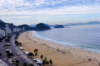 A bustling beach scene with a wide sandy shore lined by high-rise buildings. Numerous people are scattered along the beach, some under umbrellas, while others walk near the shoreline. The ocean stretches to the horizon, meeting a partly cloudy sky with a range of mountains in the distance. Palm trees and a busy road run parallel to the beach.