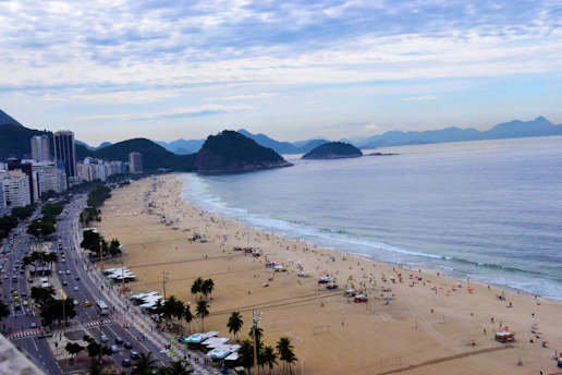 A bustling beach scene with a wide sandy shore lined by high-rise buildings. Numerous people are scattered along the beach, some under umbrellas, while others walk near the shoreline. The ocean stretches to the horizon, meeting a partly cloudy sky with a range of mountains in the distance. Palm trees and a busy road run parallel to the beach.