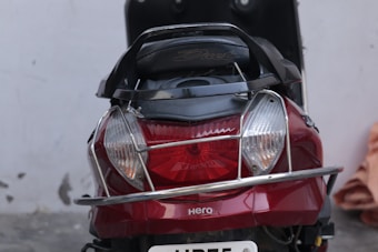 A close-up view of the rear end of a red scooter with silver accents. The scooter has a chrome protective guard around its tail light and indicators. The brand name 'Hero' is visible, along with the model name 'Duet.' There is a license plate partially showing at the bottom and a blurred background with a wall and some objects.