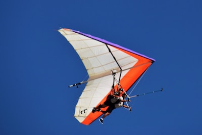 A hang glider with vibrant orange and purple accents soars against a clear blue sky. The pilot and a passenger are securely strapped in the harness, wearing helmets for safety.