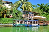 A picturesque lakeside scene with vibrant, lush greenery surrounding a wooden cabin with large windows. Two boats are moored in the bright green water in the foreground, near the cabin's dock which has large umbrellas providing shade. The backdrop features tall palm trees and dense foliage, creating a tropical atmosphere.