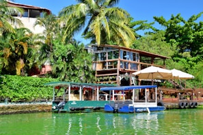 A picturesque lakeside scene with vibrant, lush greenery surrounding a wooden cabin with large windows. Two boats are moored in the bright green water in the foreground, near the cabin's dock which has large umbrellas providing shade. The backdrop features tall palm trees and dense foliage, creating a tropical atmosphere.