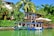 A picturesque lakeside scene with vibrant, lush greenery surrounding a wooden cabin with large windows. Two boats are moored in the bright green water in the foreground, near the cabin's dock which has large umbrellas providing shade. The backdrop features tall palm trees and dense foliage, creating a tropical atmosphere.