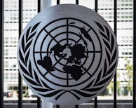 A metal emblem consisting of a world map surrounded by two olive branches is mounted on a black barred fence. The background shows a blurred view of a modern building with large rectangular windows.
