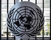 A metal emblem consisting of a world map surrounded by two olive branches is mounted on a black barred fence. The background shows a blurred view of a modern building with large rectangular windows.