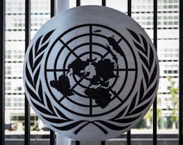 A metal emblem consisting of a world map surrounded by two olive branches is mounted on a black barred fence. The background shows a blurred view of a modern building with large rectangular windows.