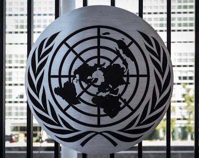 A metal emblem consisting of a world map surrounded by two olive branches is mounted on a black barred fence. The background shows a blurred view of a modern building with large rectangular windows.