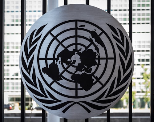 A metal emblem consisting of a world map surrounded by two olive branches is mounted on a black barred fence. The background shows a blurred view of a modern building with large rectangular windows.