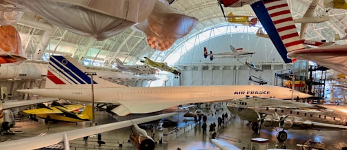 A large aviation museum exhibit featuring multiple aircraft. The main focus is a Concorde jet with Air France livery, surrounded by other historic airplanes both on the ground and suspended from the ceiling. The setting includes high ceilings with natural light streaming in through large windows.