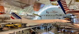 A large aviation museum exhibit featuring multiple aircraft. The main focus is a Concorde jet with Air France livery, surrounded by other historic airplanes both on the ground and suspended from the ceiling. The setting includes high ceilings with natural light streaming in through large windows.