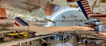A large aviation museum exhibit featuring multiple aircraft. The main focus is a Concorde jet with Air France livery, surrounded by other historic airplanes both on the ground and suspended from the ceiling. The setting includes high ceilings with natural light streaming in through large windows.